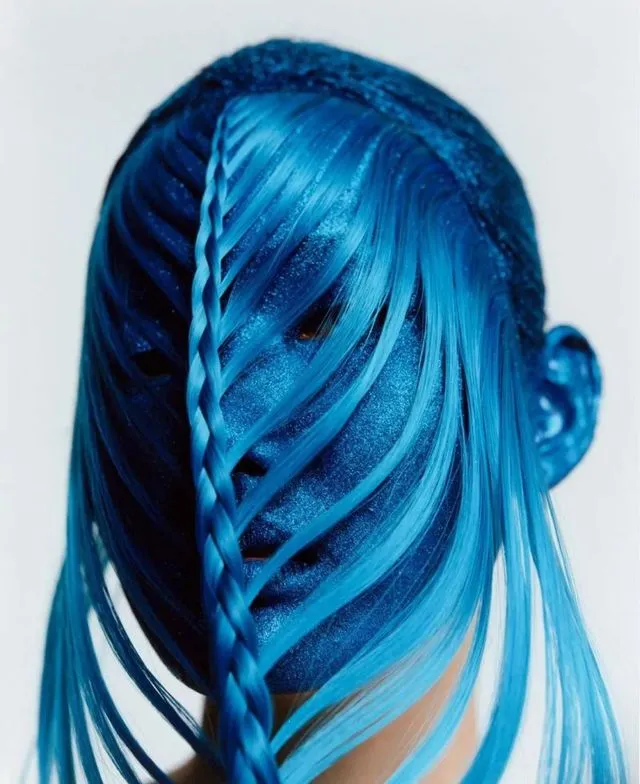 Close portrait of a face covered in blue pigment, with a braid and loose blue hair crossing the features. Photography by Cho Gi-Seok.